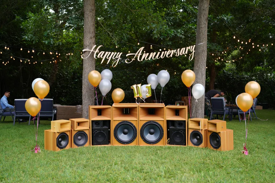 Décor de fête en extérieur avec haut-parleurs DIY en bois, ballons dorés et argentés, guirlande lumineuse “Happy Anniversary” et chiffres 11.1 sur le podium audio.